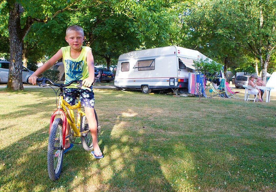 Child on a bike in front of a caravan set up on a shady campsite pitch, enjoying a summer family atmosphere.