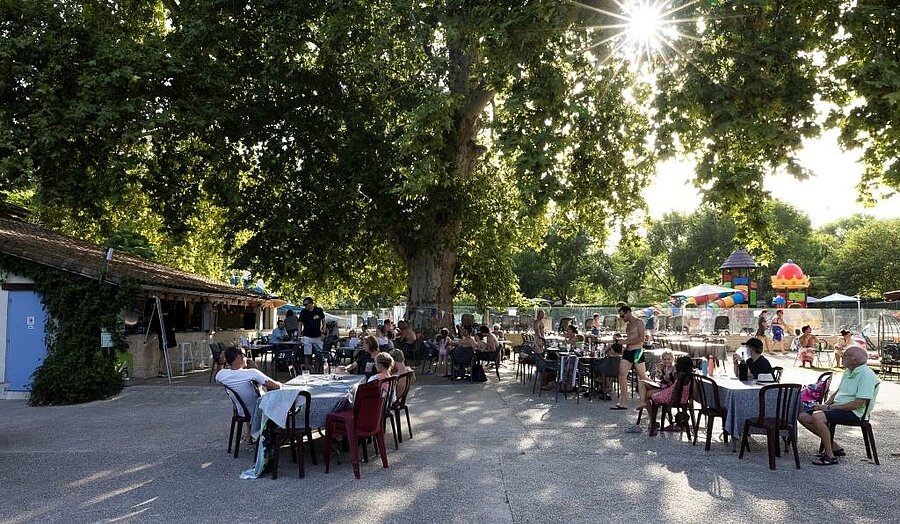 Guests relaxing in the shaded bar-restaurant area of Le Moulin campsite near Toulouse, with kids’ play area in the background.