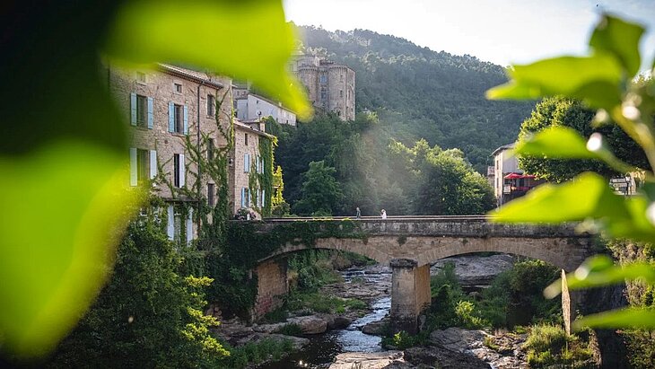 View of Largentière Bridge, stone houses, and a medieval tower, surrounded by greenery along the riverbank.