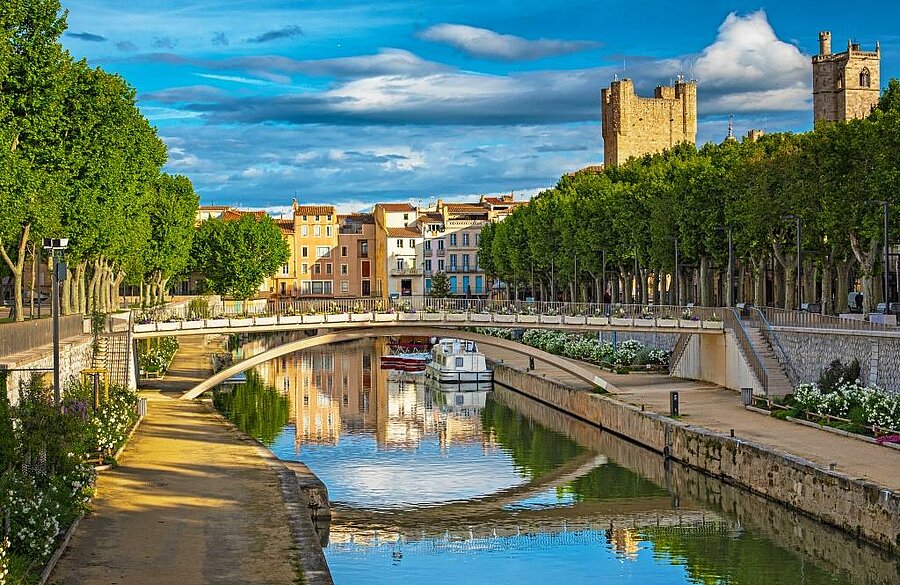 View of the Robine Canal in Narbonne, lined with trees and overlooked by the medieval tower, close to a nature campsite.