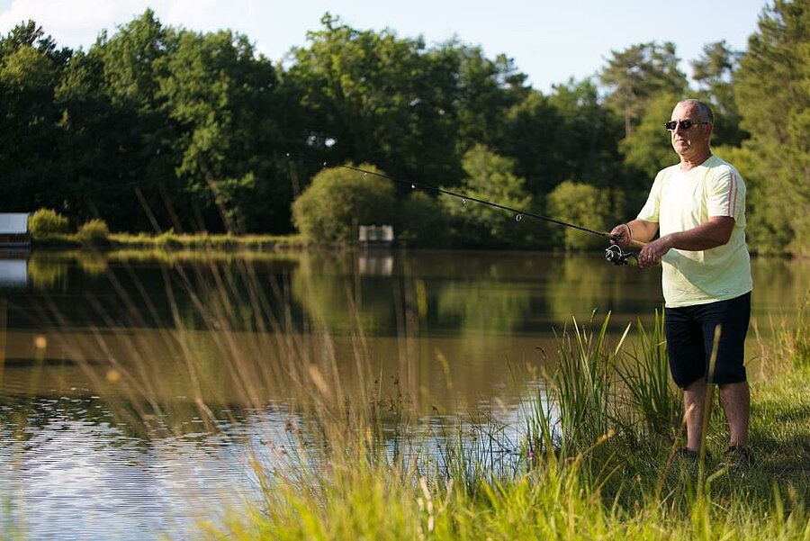 Angler casting his line by a pond at a fishing campsite, green natural setting with a peaceful atmosphere.