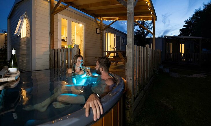 Couple relaxing in a private spa beside a lit wooden accommodation, cosy setting illustrating an early booking couple.