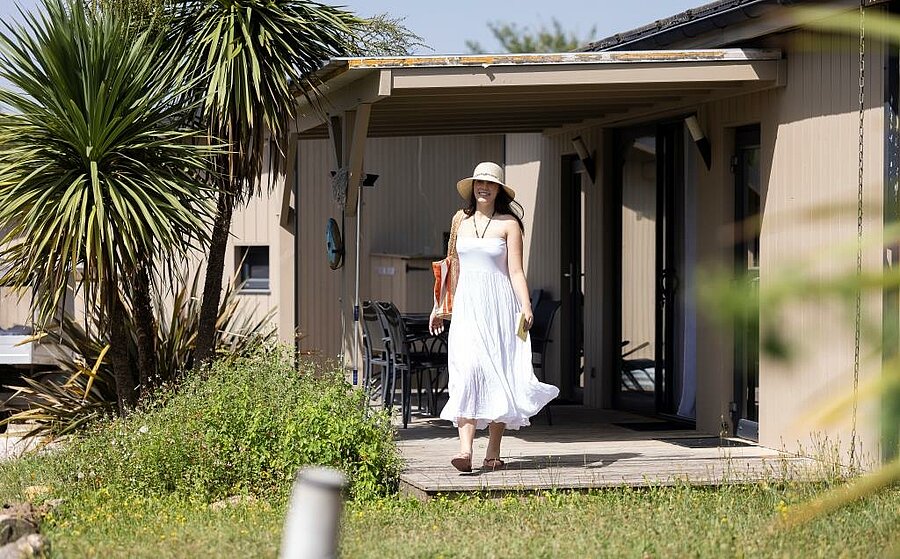 Woman in a white dress stepping out of a modern chalet with shaded terrace, in a nature campsite near Toulouse.