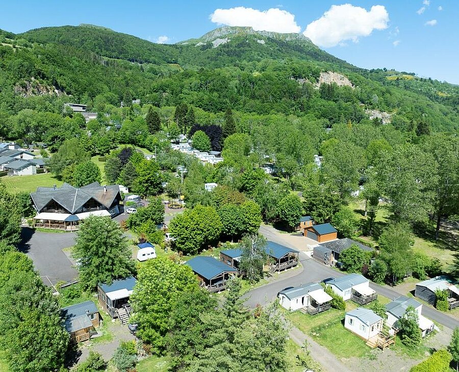 Aerial view of a green campsite in Auvergne with mobile homes, chalets, and a forest in the background.