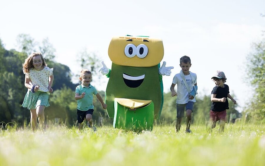 Children running through a lush green field alongside PAKO, a cheerful mascot, during a family activity at a campsite near Honfleur, in Normandy.