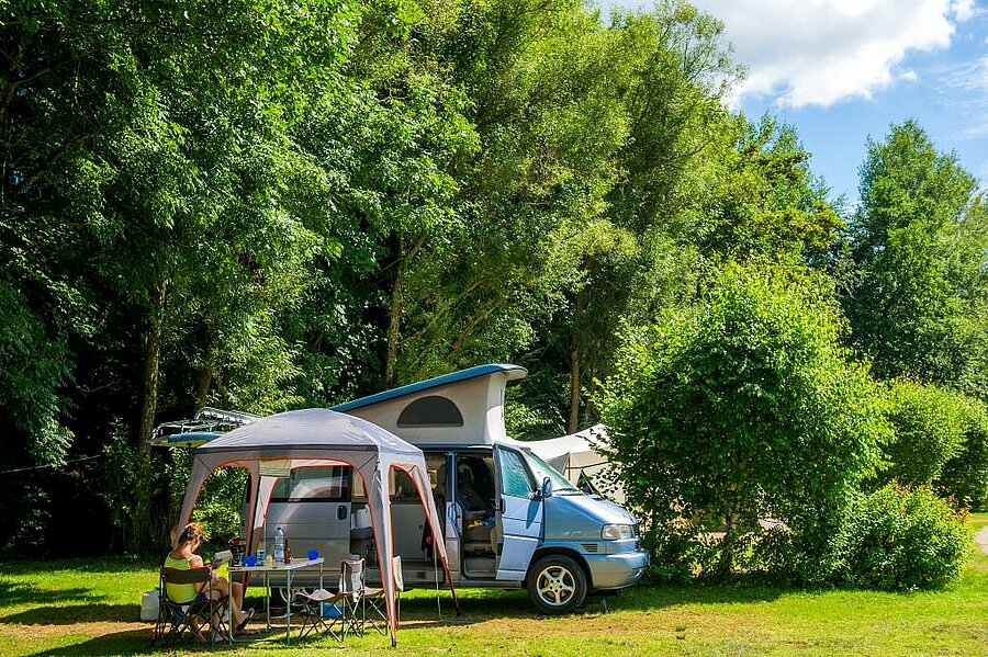 Camper van set up on a spacious, shaded van pitch, with outdoor dining area in lush natural surroundings.