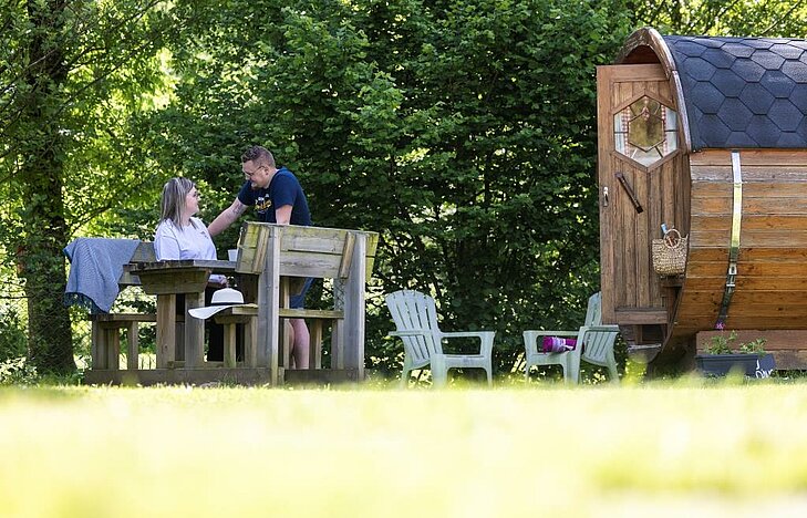 A couple chatting near an unusual barrel accommodation, set in a lush green environment for a peaceful getaway for two.