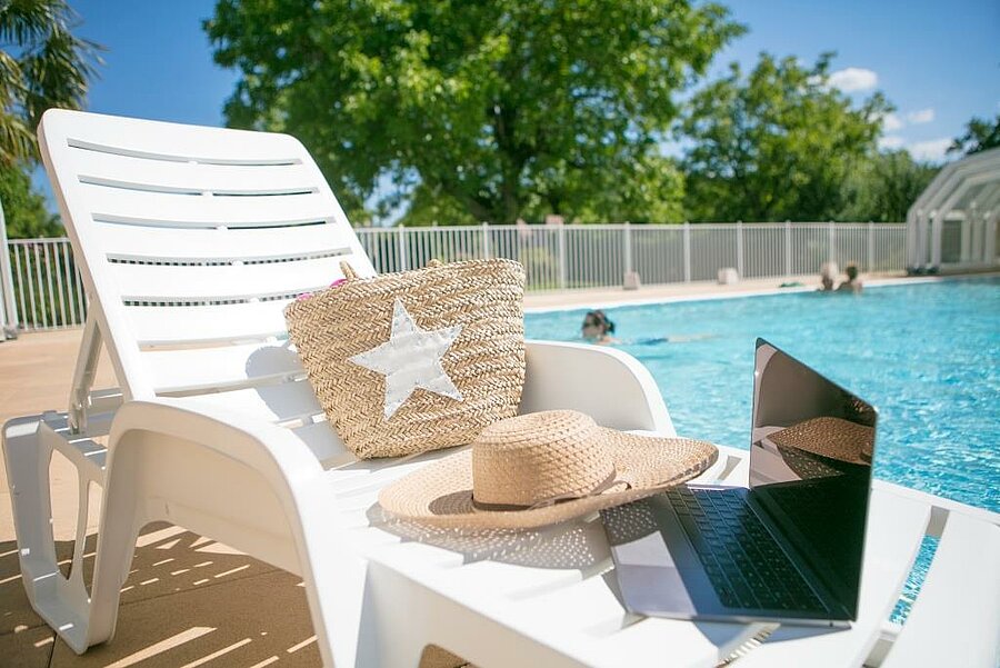 Sunlounger by the pool with hat, basket and laptop – the picture of a relaxing, connected break at a budget-friendly campsite with a best price guaranteed.