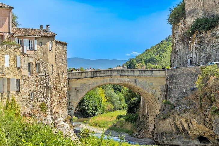 Roman bridge in Vaison-la-Romaine spanning the Ouvèze River, with historic houses, a cliff and hills in the background.