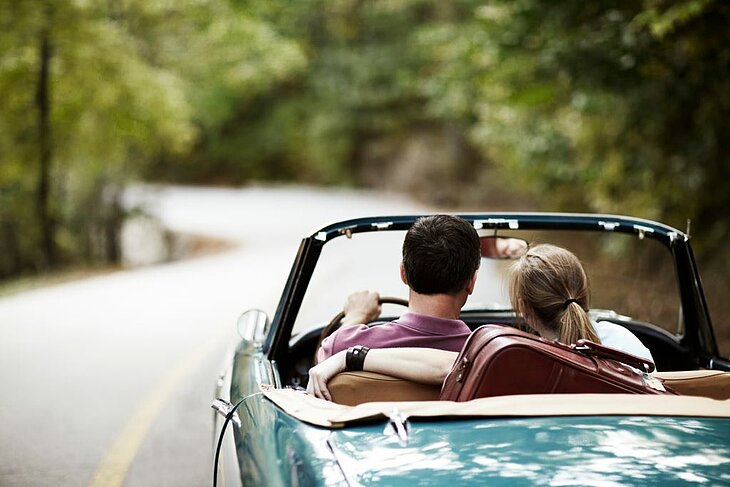 A couple in a convertible driving along a nature-lined road, a symbol of freedom and looking ahead for a getaway for two.