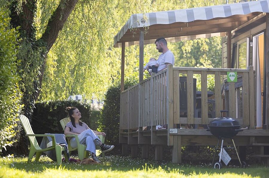 [Translate to Anglais:] Famille profitant d’un moment de détente sur la terrasse d’un mobil-home ombragé, dans un camping avec parc aquatique et cadre naturel verdoyant