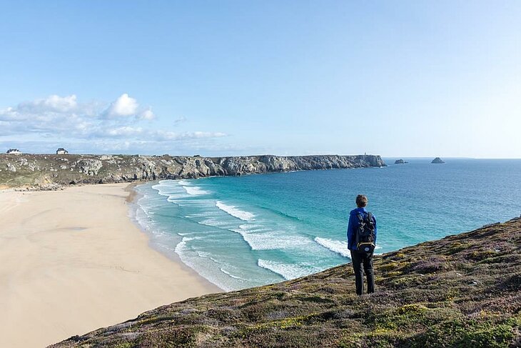 Hiking overlooking the Breton ocean Hiker gazing over a vast beach and the ocean from the wild coast of Finistère, an iconic nature activity of Finistère Tourism.