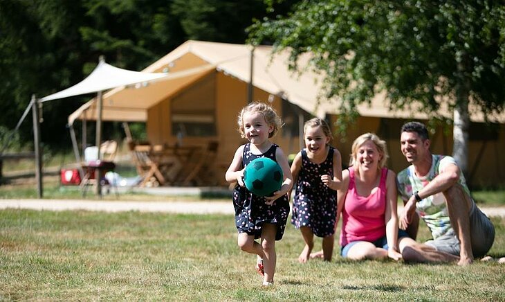 Little girl playing with a ball in front of her smiling family at a nature campsite, symbolizing the simple joy of early holiday moments.
