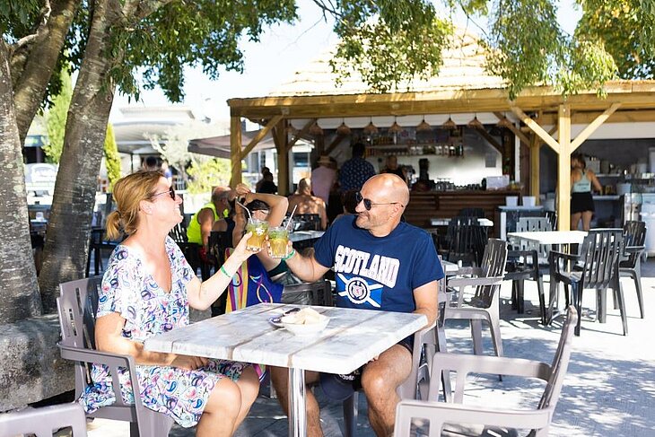 Couple toasting on a shaded campsite bar terrace, friendly moment illustrating an early booking couple.
