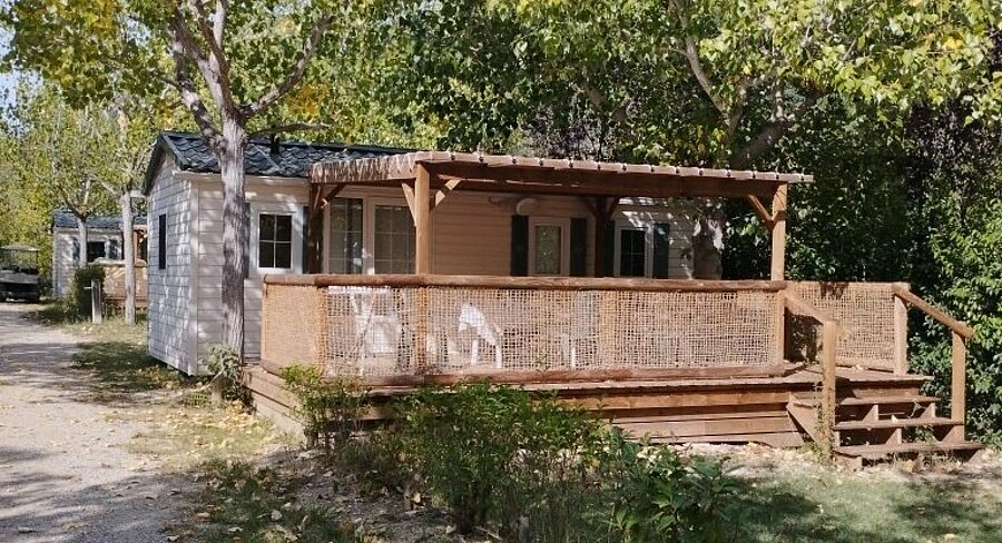 Mobile home with a covered wooden terrace and outdoor furniture, surrounded by trees at Domaine de l’Écluse in Drômes.