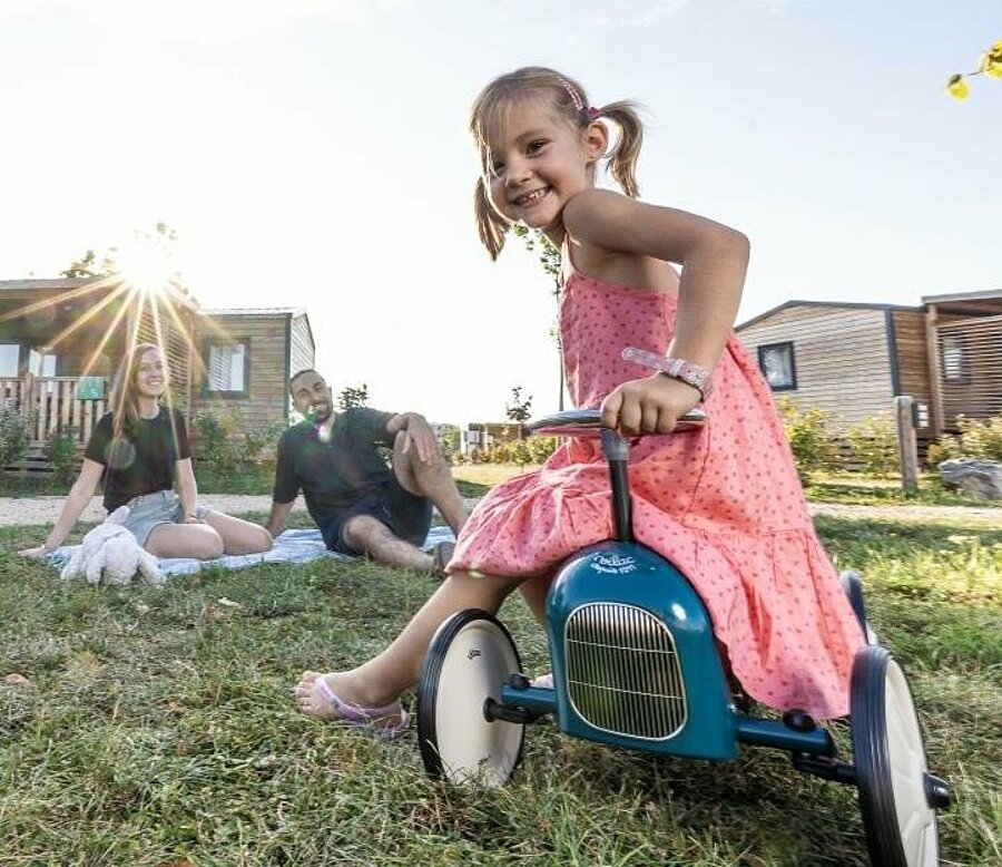Smiling little girl on a balance bike in front of a mobile home, her parents relaxing in the sun — the perfect image of a family nature getaway with the Campsite Early Booking offer “A Little Happiness Ahead”.