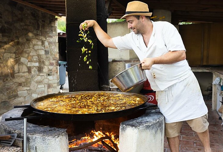 Julien preparing his famous wood-fired paella at the La Pinède campsite restaurant, a friendly and iconic moment of tourism in Lézignan-Corbières, blending family recipes with produce from the vegetable garden.