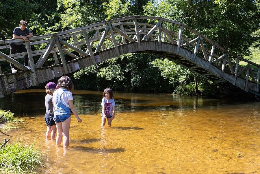 [Translate to Anglais:] Enfants jouant dans une rivière peu profonde sous un pont en bois, dans un camping pas cher et nature idéal pour des vacances familiales au bord de l'eau