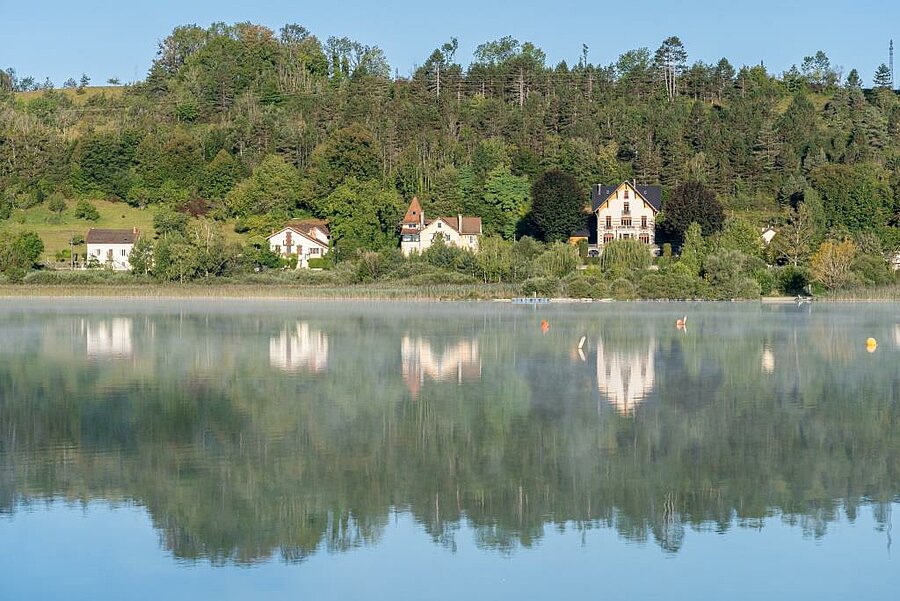 Houses by Clairvaux-les-Lacs lake with reflections on the water, discover Jura nature and heritage.