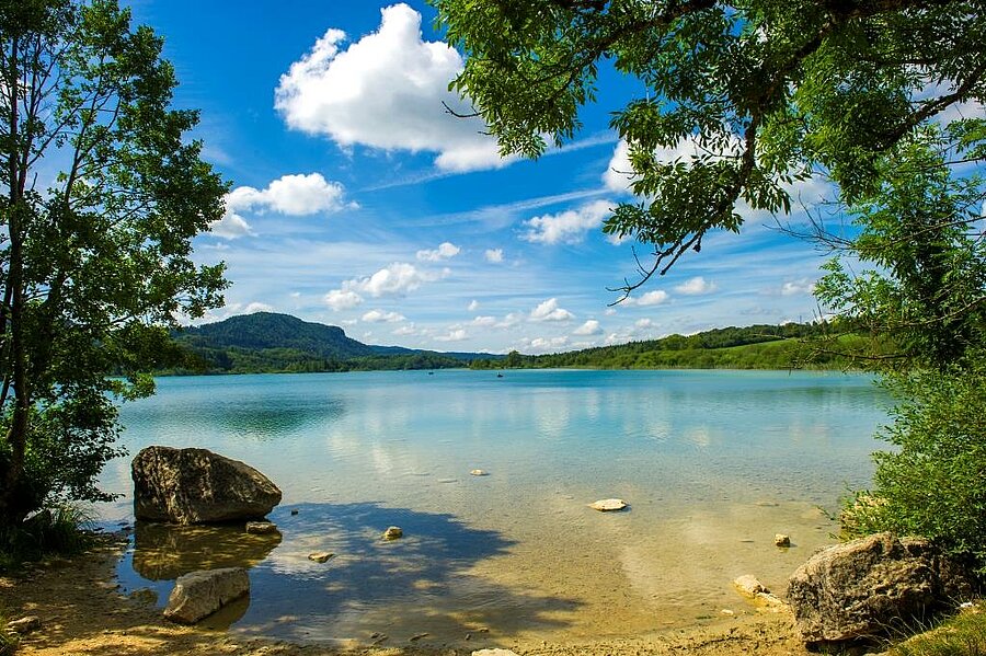 Picturesque view of a lake in the Jura region of France, surrounded by greenery and a blue sky, perfect for activities like kayaking and hiking during a camping stay in North-East France.