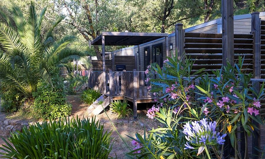 Contemporary mobile home with shaded terrace, surrounded by palm trees and Mediterranean flowers in a campsite near Nice.