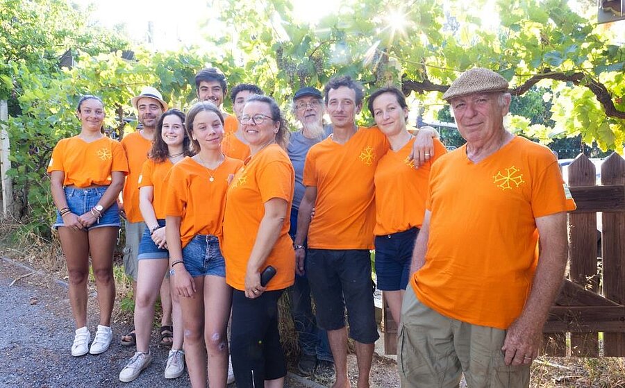 Smiling team of La Pinède campsite near Carcassonne, wearing orange t-shirts under a vineyard pergola.