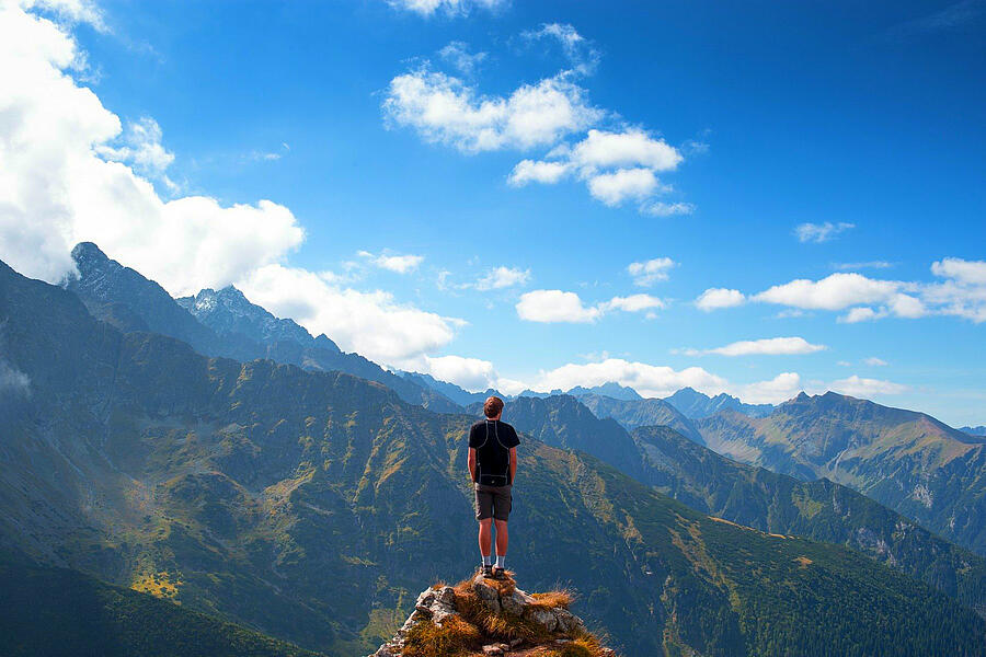 Hiker standing on a mountain peak with panoramic views of the Midi-Pyrénées mountain range under a partly cloudy blue sky.