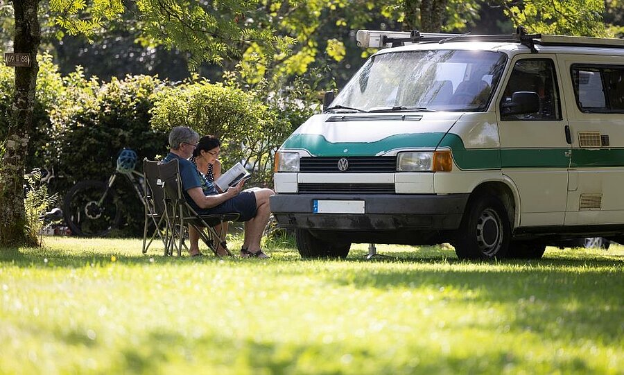 A couple relaxing in front of their campervan on a leafy pitch, capturing the freedom and simplicity of vanlife surrounded by nature.