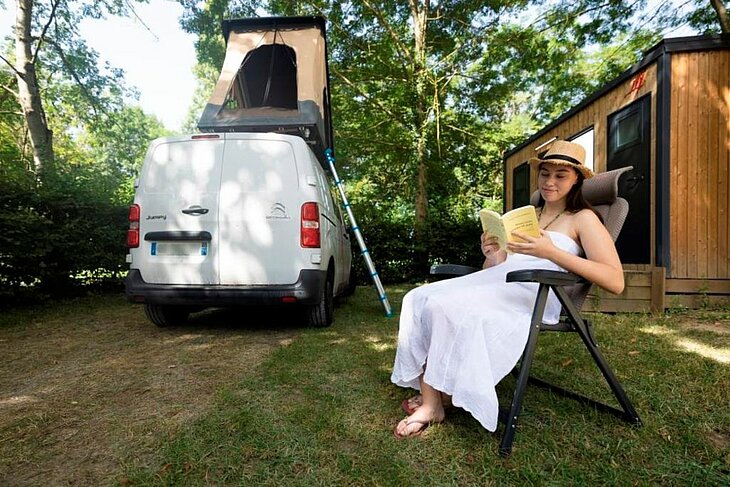 A holidaymaker reading in the shade beside a camper van with a pop-up roof at a campsite.