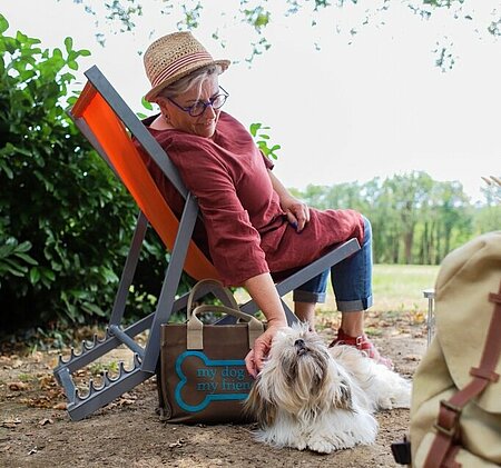 A relaxed woman in a deck chair pets her dog at a small-scale campsite where animals are welcome.