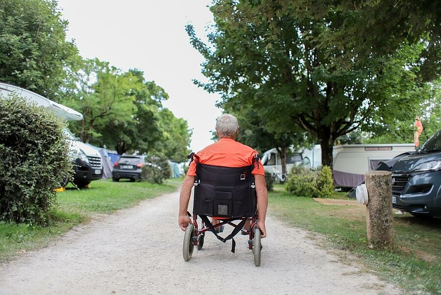 Guest in a wheelchair moving along a shaded campsite path, showing an accessible mobile home with comfortable and adapted facilities.
