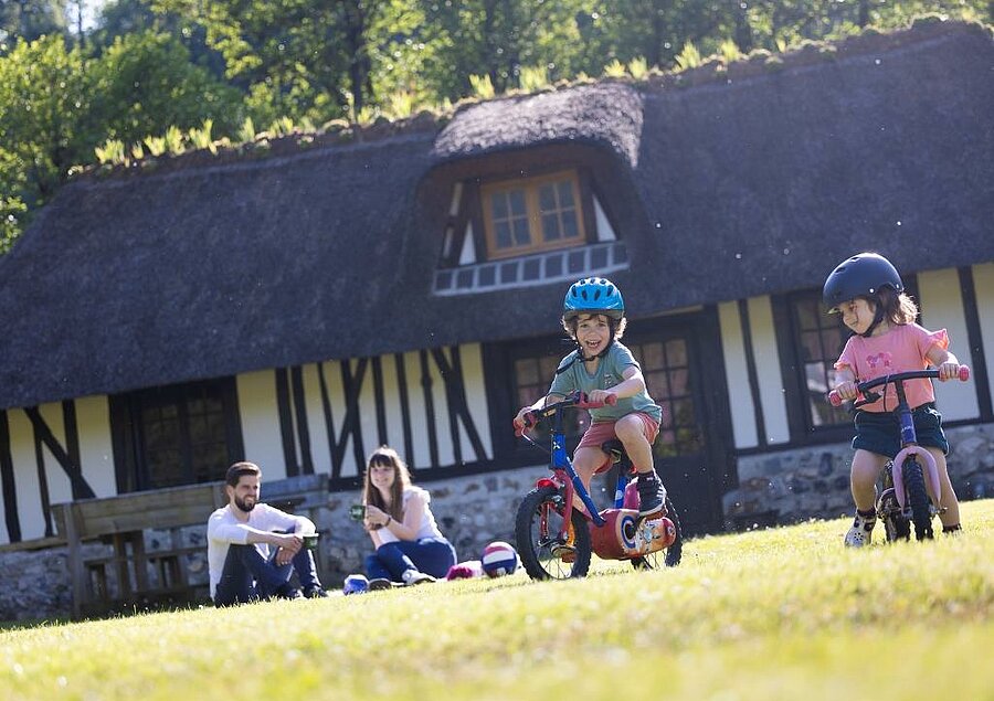 Two children riding their bikes in front of a traditional Norman accommodation, while their parents enjoy a relaxing moment at a campsite near Honfleur.
