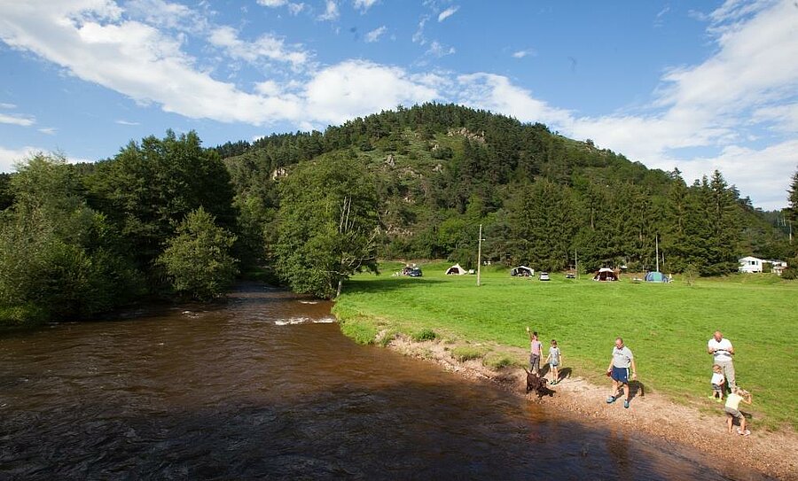 Family walking by a river near a lakeside campsite, green pitches and forested hills in the background.