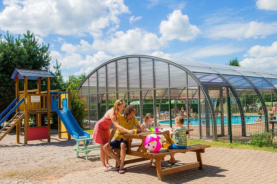 A family enjoying a friendly moment near the covered pool and playground at a Sites et Paysages campsite during a sunny Ascension weekend.