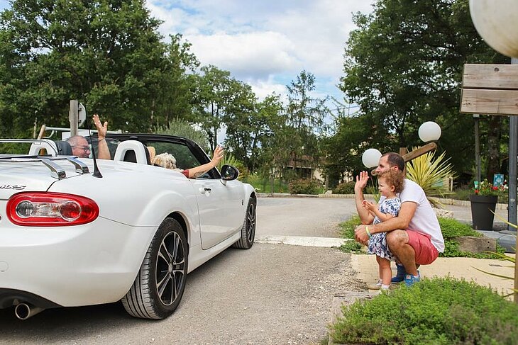 Family and senior couple waving at each other at a campsite entrance, symbolizing friendliness and the joy of simple holidays.